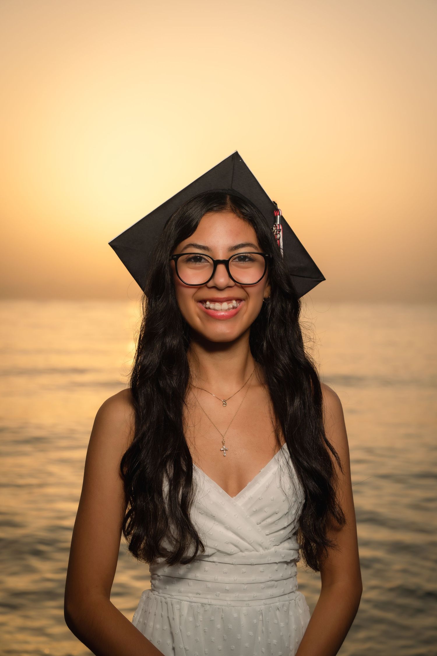 A graduate poses in a white dress against a sunset backdrop over the ocean.
