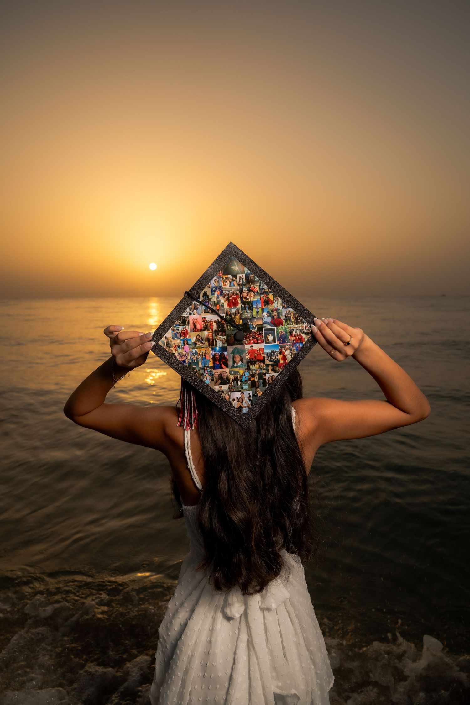 A graduation cap featuring colorful decorated artwork is shown against a dramatic sunset over the ocean.