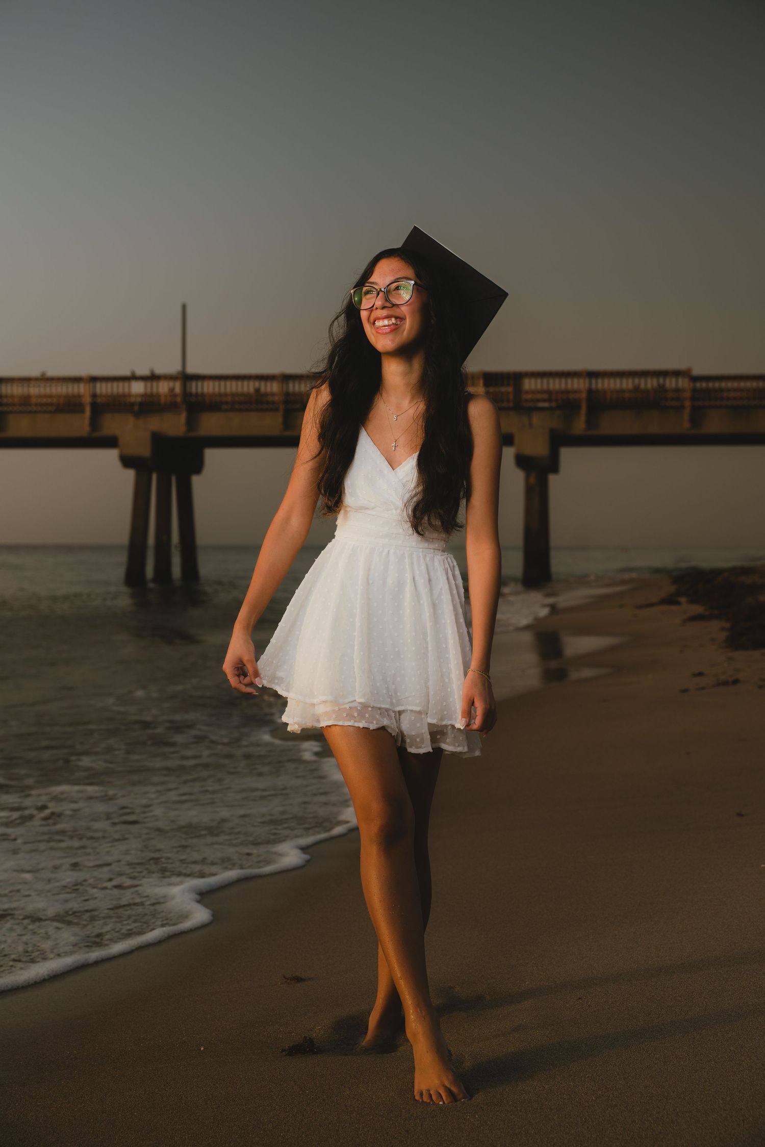 A figure in white dress and graduation cap walks barefoot along a beach pier at sunset.