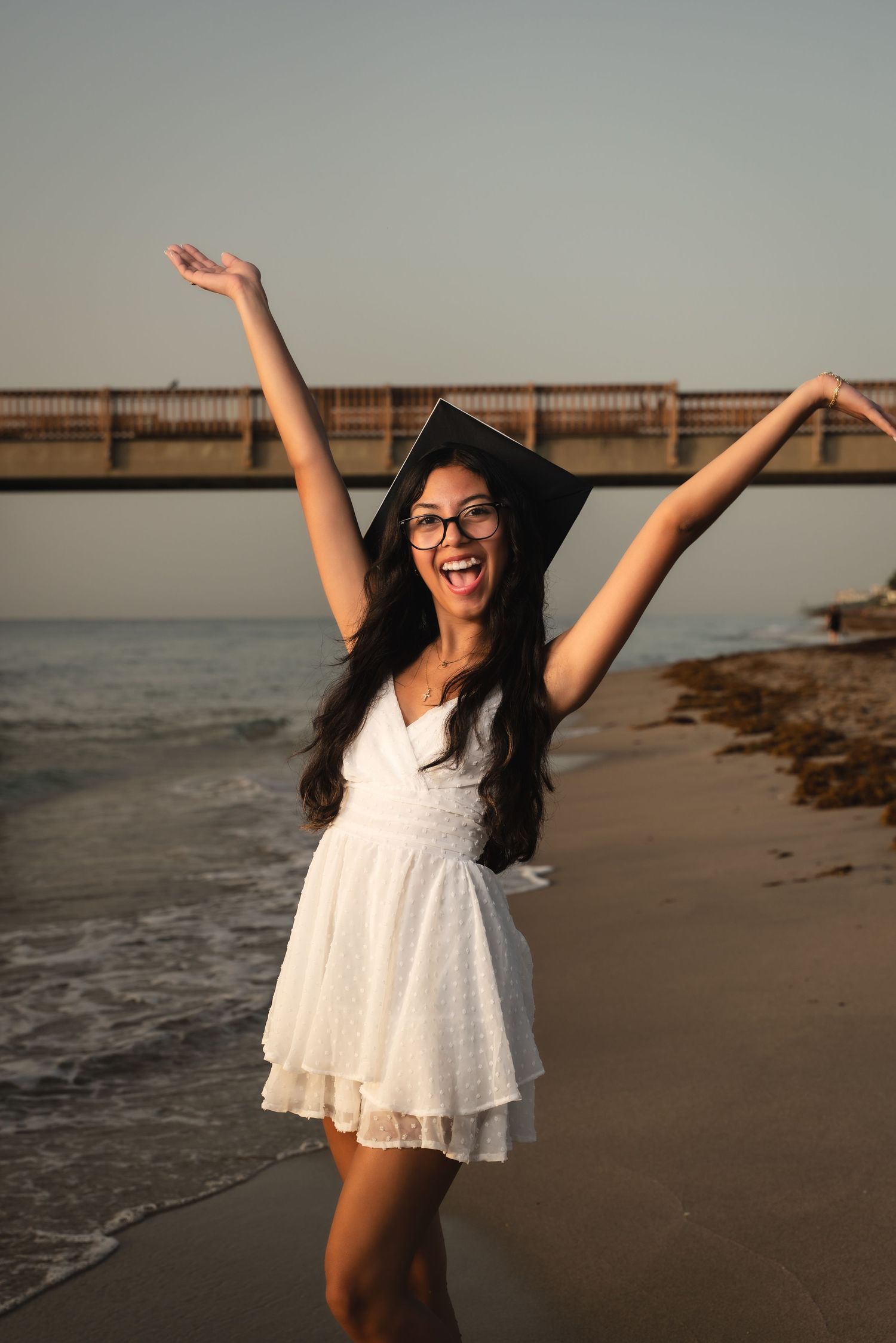 A graduate in a white dress celebrates on the beach with arms raised in joyful expression.