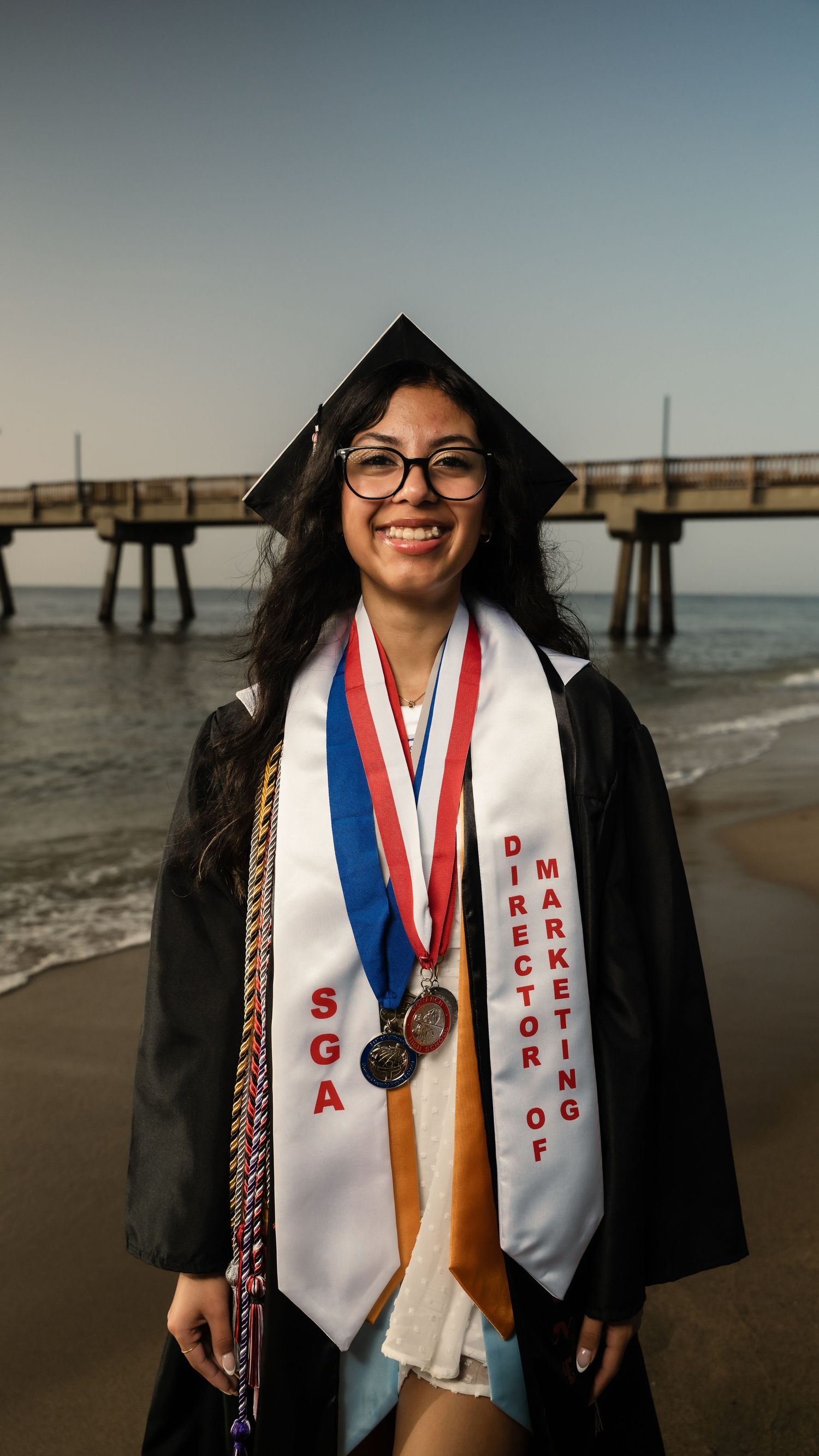 Graduate in cap and gown stands on beach with graduation stole and honor cords near pier at sunset.