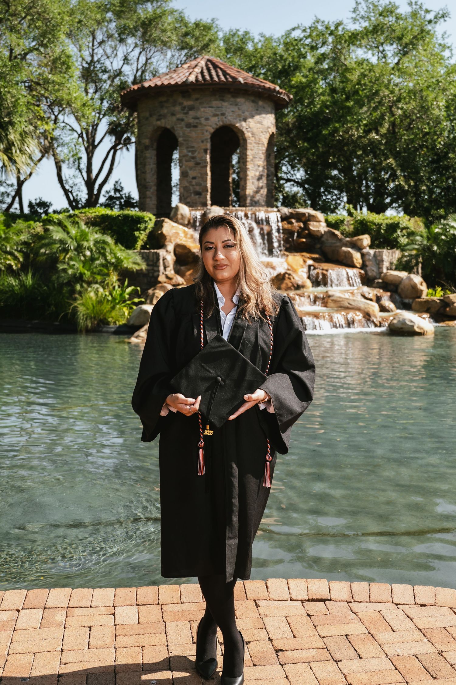 Standing by a garden gazebo and waterfall feature with brick pavers and lush greenery in the background.