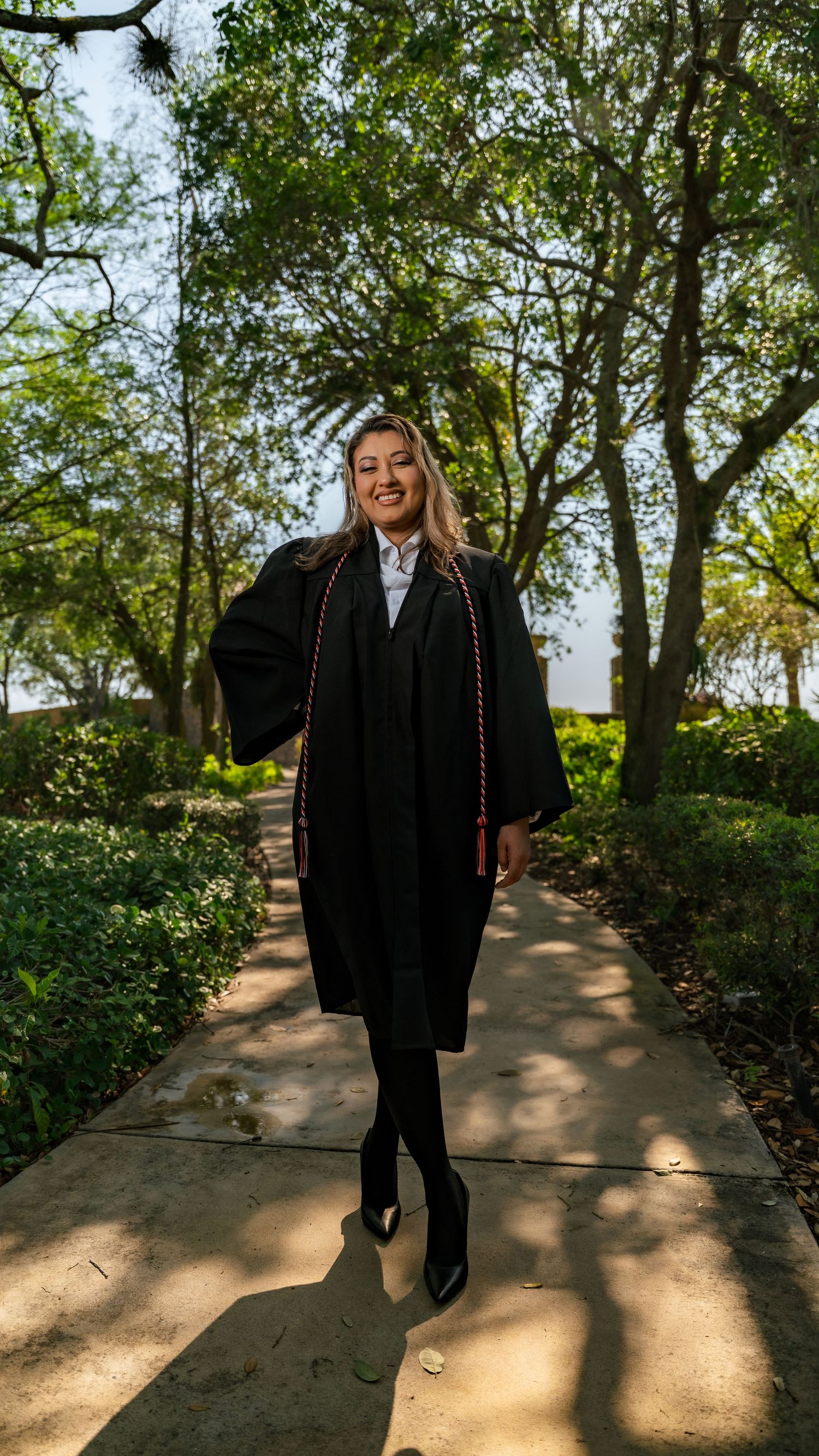 A person wearing all black clothing walks on a sunlit garden path surrounded by lush green trees and landscaping.