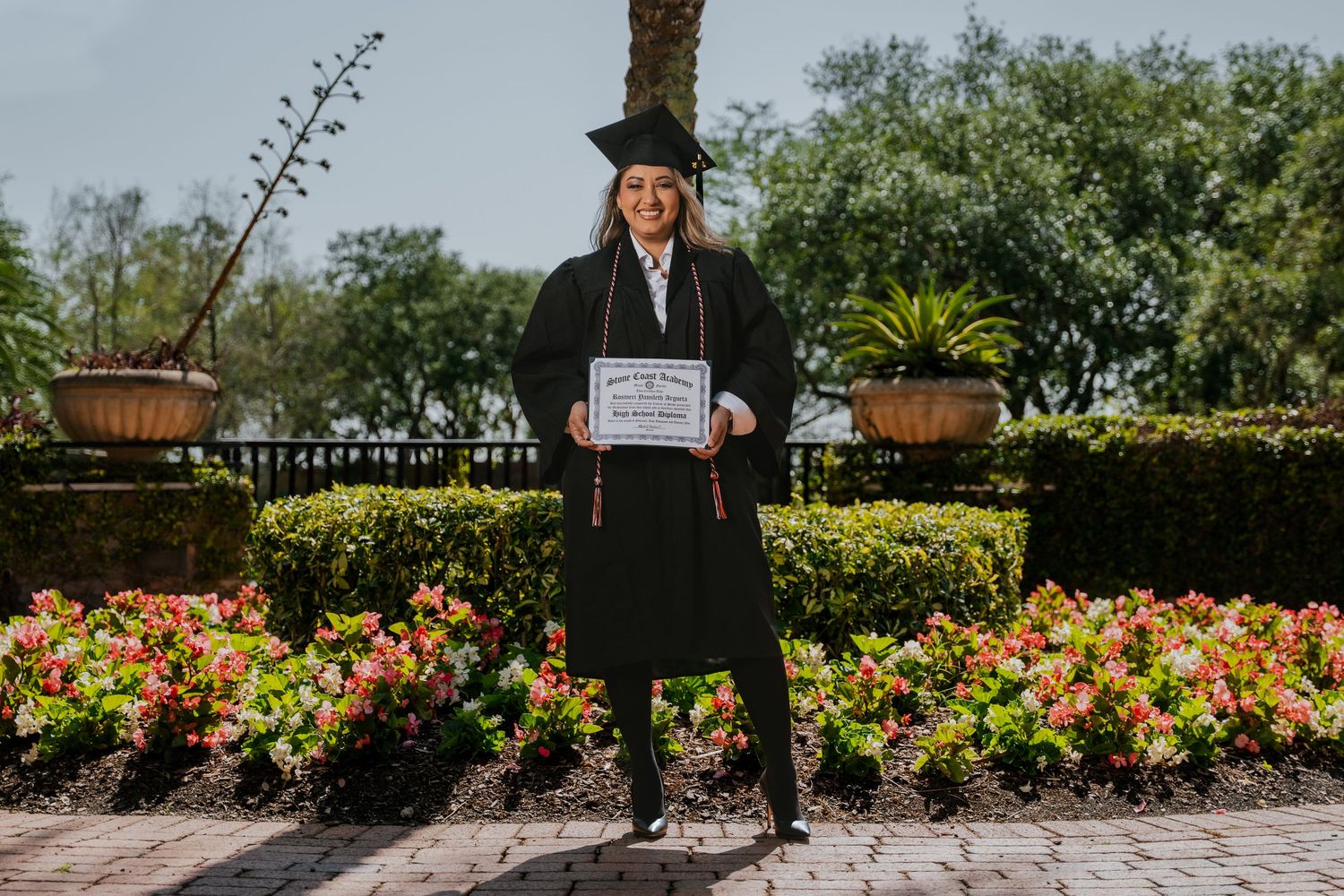 Graduate in cap and gown poses among colorful flower gardens in landscaped outdoor setting.