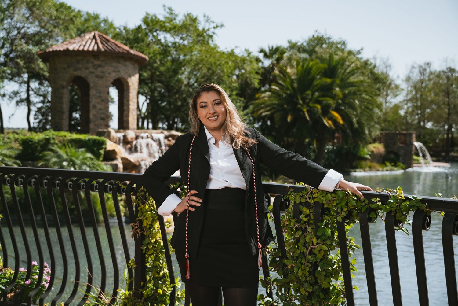 A person in business attire poses on a decorative bridge railing near a gazebo and palm trees in a tropical garden setting.