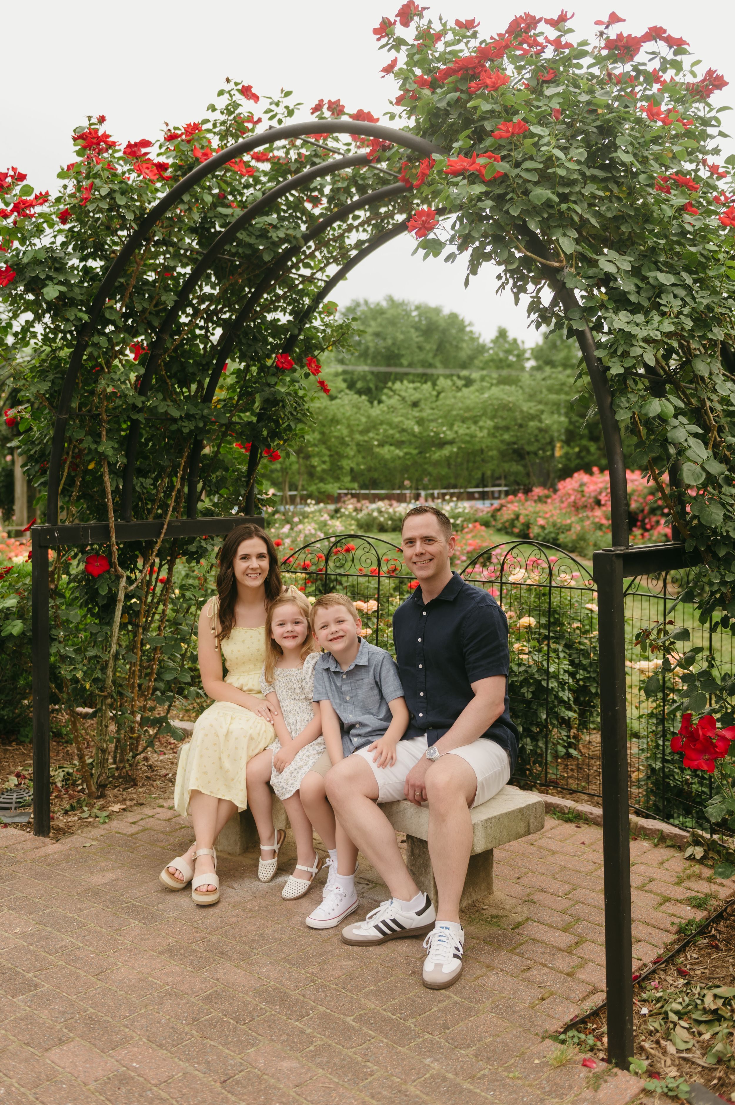 Family of four smiling on a bench under a red rose garden arch.
