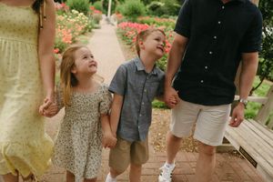 Family with two young children walking hand-in-hand along a brick garden path surrounded by colorful flowers.