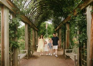 Family of four walks under a lush green garden pergola tunnel, parents and two children in coordinating outfits.