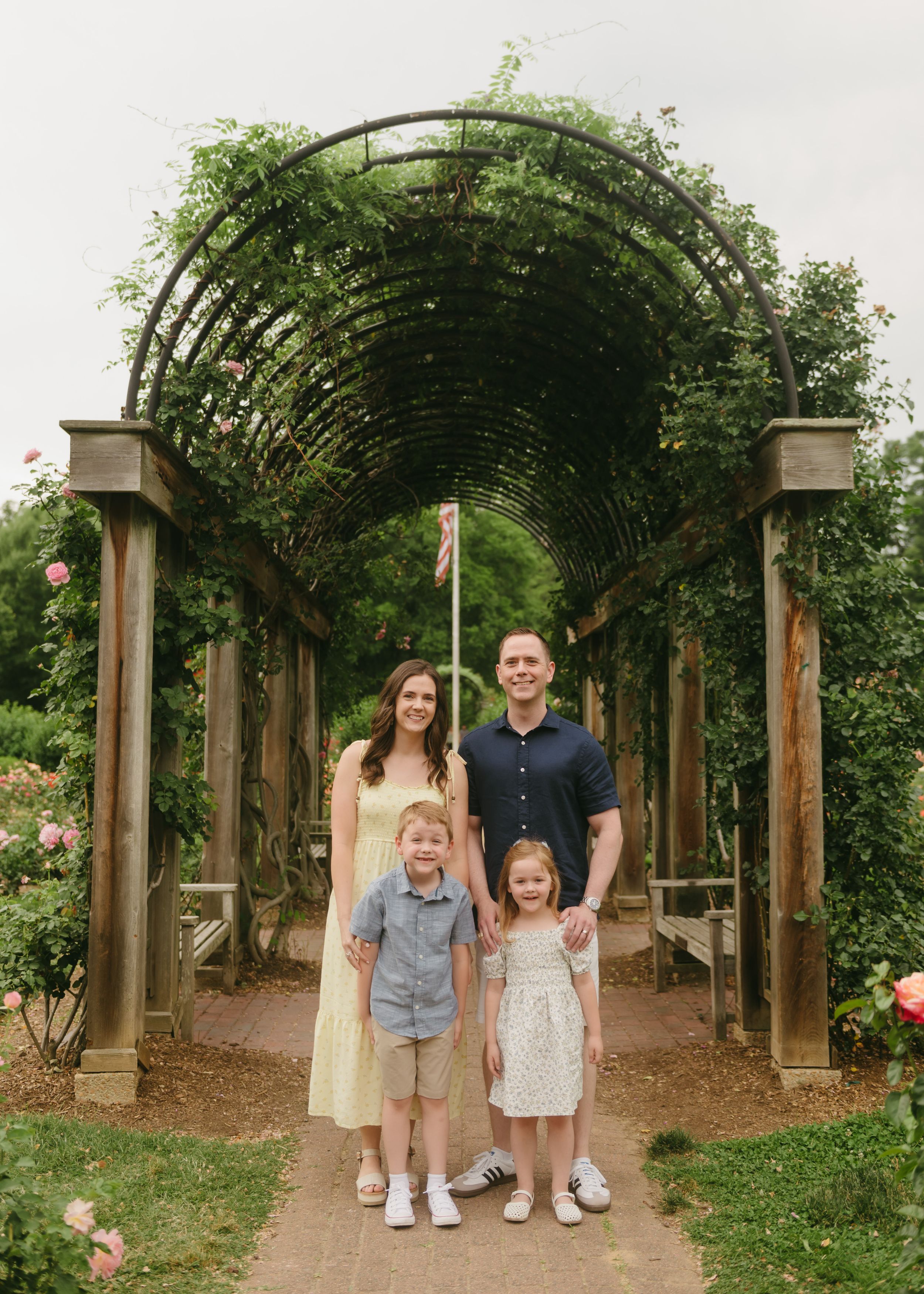 Family of four posing under a lush garden archway surrounded by roses and greenery on a cloudy day.