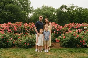 Family of four posing in front of vibrant pink rose bushes in a lush garden setting.