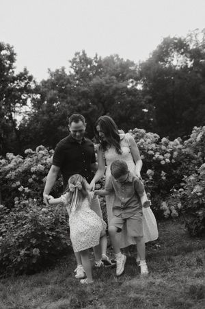 Black and white family photo of parents and two young children holding hands in a lush garden.