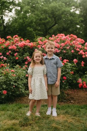 Two young siblings smiling together in front of vibrant pink rose bushes in a garden.