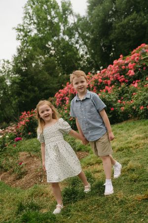 Two young children holding hands in a garden with pink roses, girl in floral dress and boy in blue shirt.