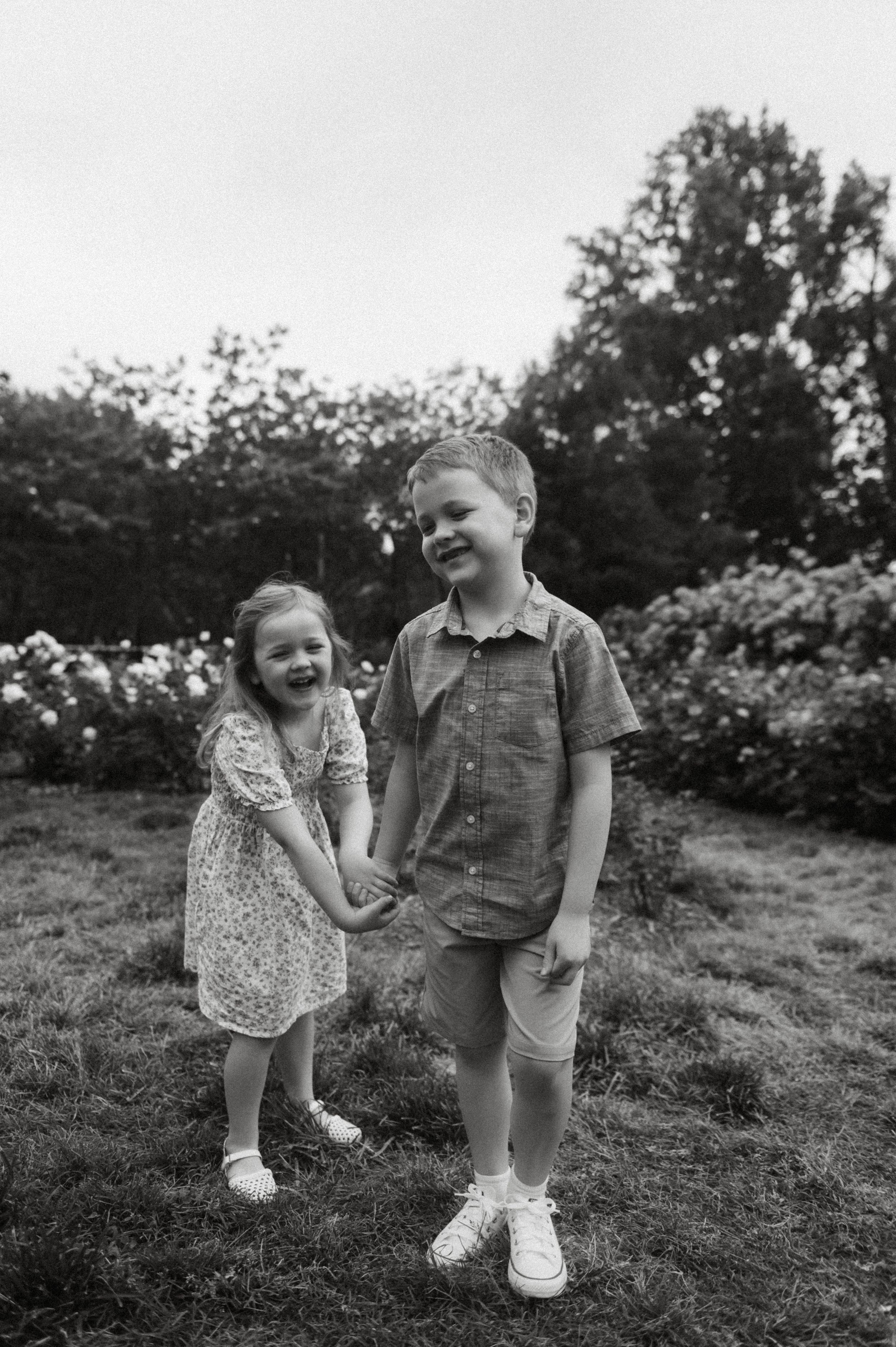 Black and white photo of two young siblings holding hands and smiling in a lush garden.