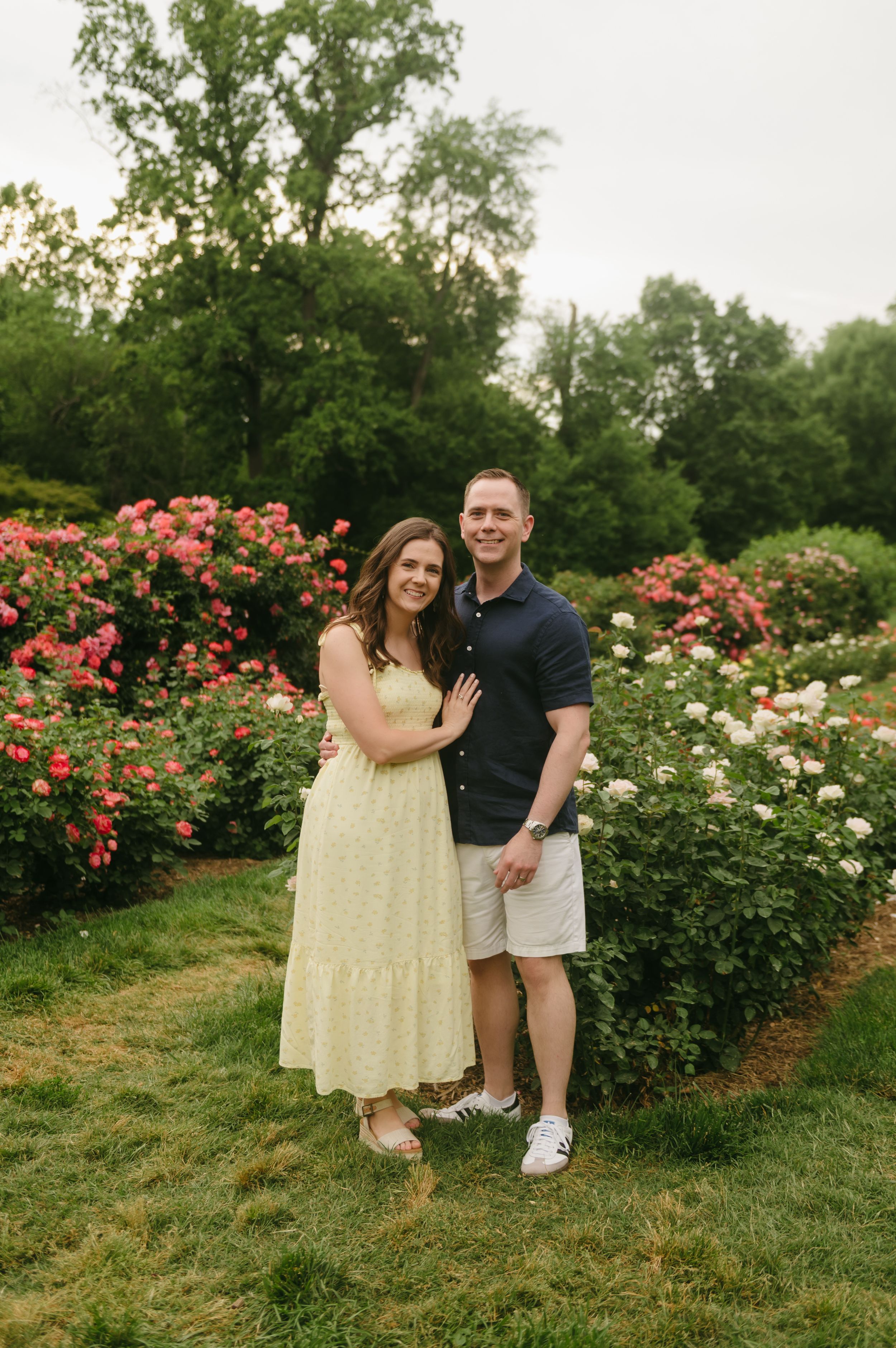 Couple smiling together in a colorful rose garden, woman in yellow dress, man in navy polo.