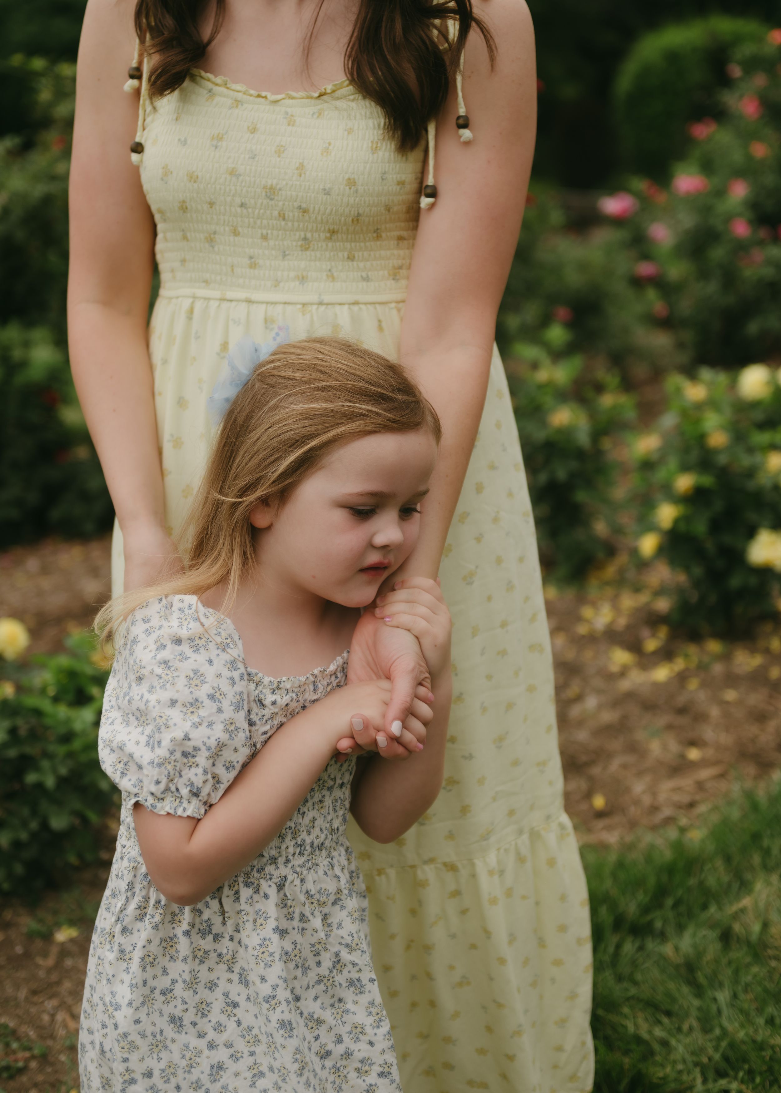 A young girl in a floral dress holds her mother's hand in a garden with colorful roses blooming in the background.