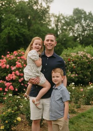 A man holds a young girl while a boy stands beside him in a lush garden with pink roses blooming.