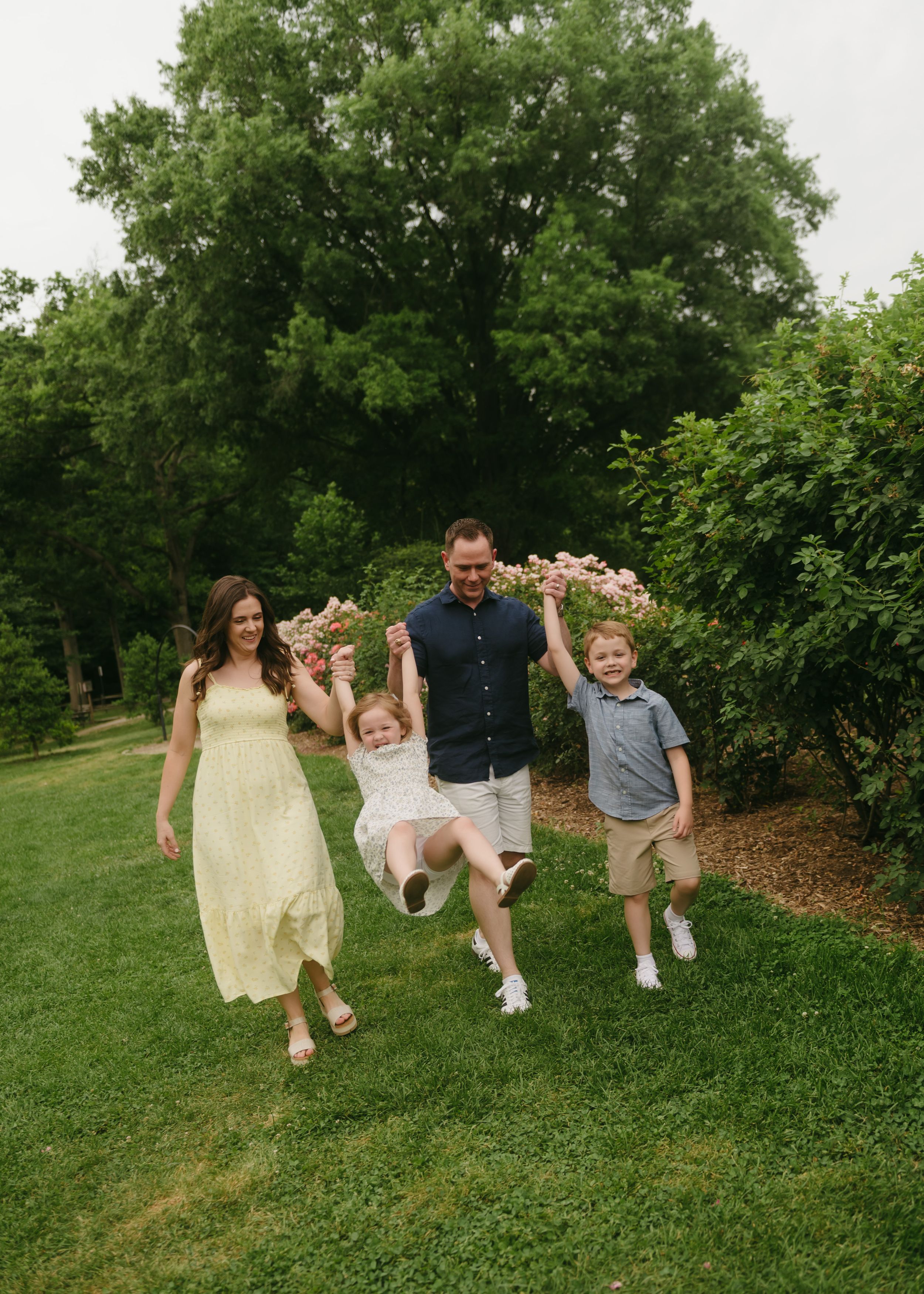 A joyful family of four plays outdoors, swinging a young girl between parents and son in a lush green garden.