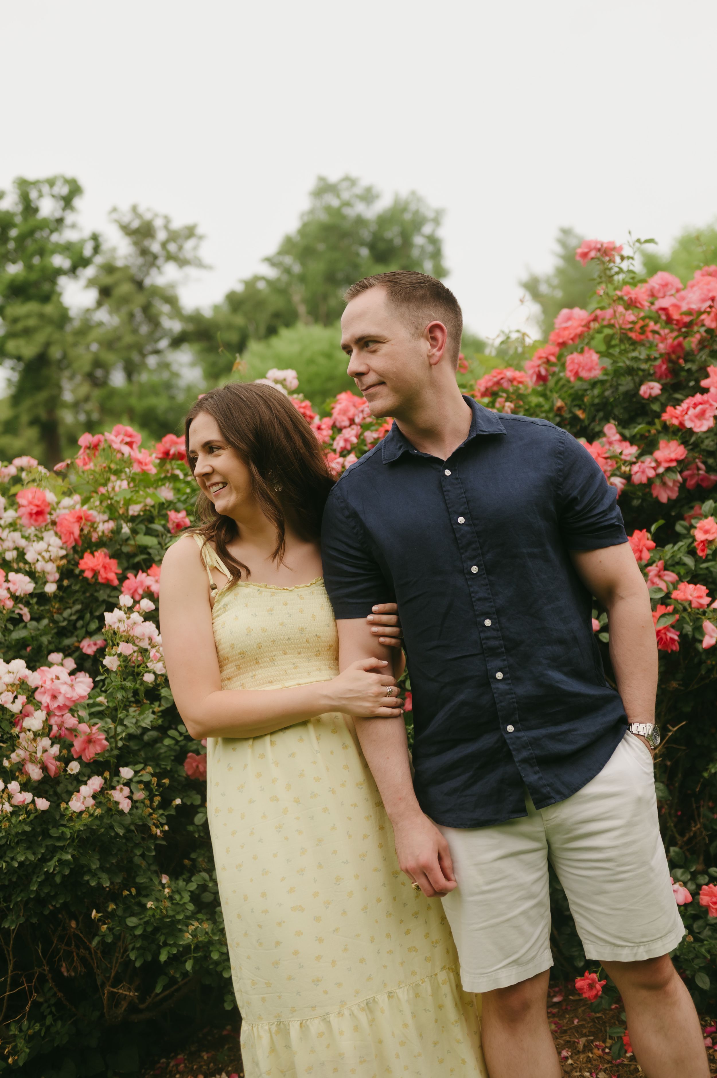 Couple smiling together in front of vibrant pink rose bushes, woman in yellow dress, man in navy shirt.