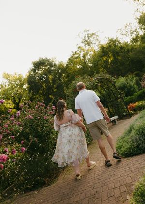 Couple walking hand-in-hand along a brick garden path surrounded by blooming roses and lush greenery at sunset.