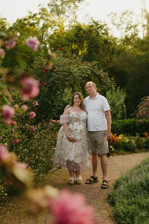 Couple poses in a lush garden path surrounded by blooming pink roses and green archway during golden hour.