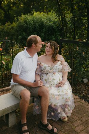 Pregnant woman in floral dress sits with partner on garden bench surrounded by lush greenery during maternity photo session.