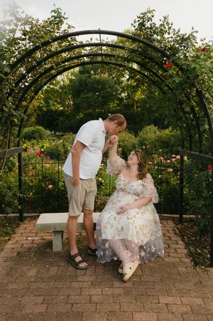 A couple shares a tender moment under a floral garden arch, woman in floral dress seated on bench, man leaning down to kiss her hand.