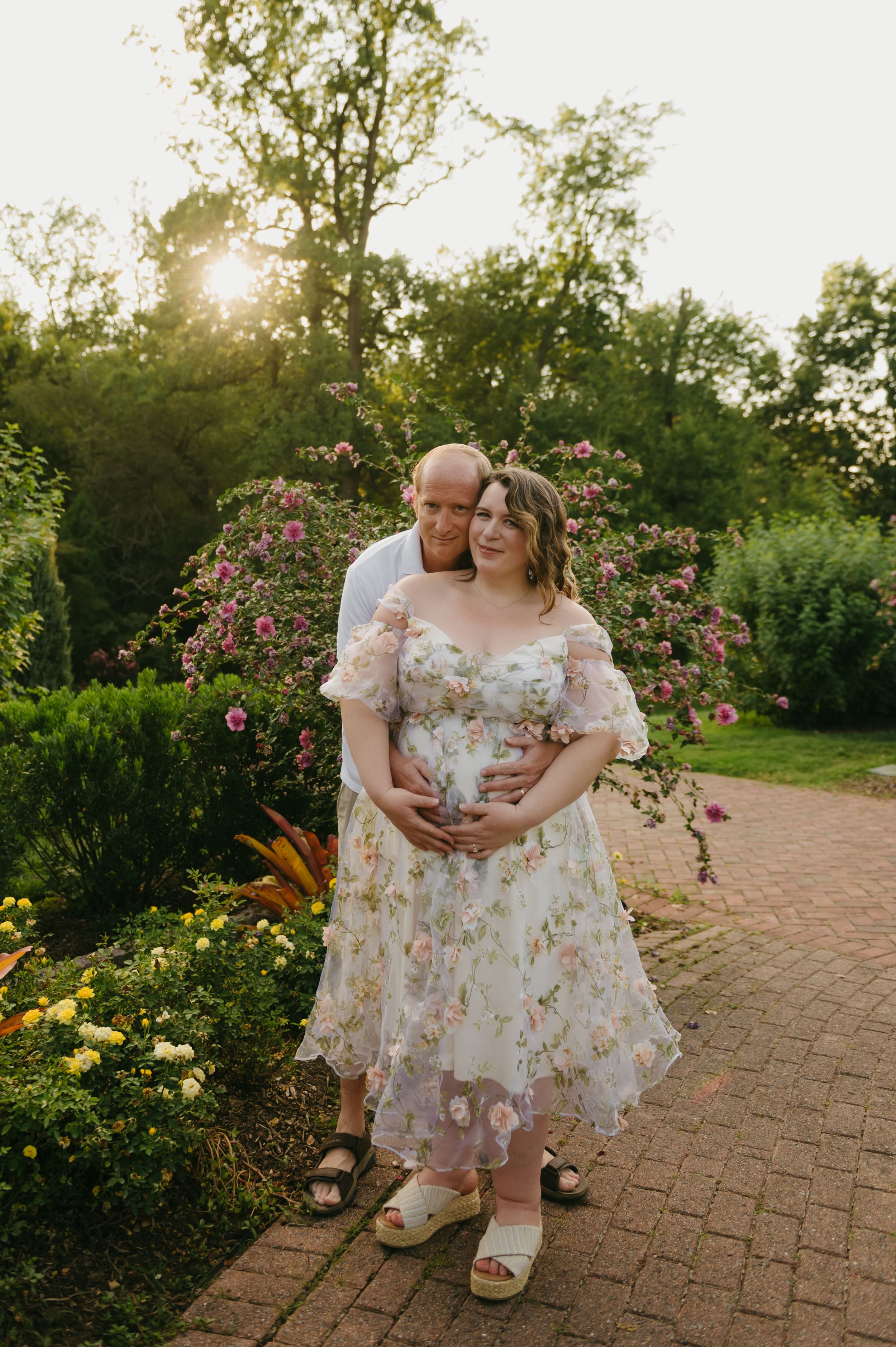 Couple poses in a blooming garden at golden hour, woman in floral dress with partner embracing her from behind on brick path.