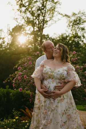 A couple shares a tender maternity moment at golden hour, surrounded by blooming pink roses in a lush garden.
