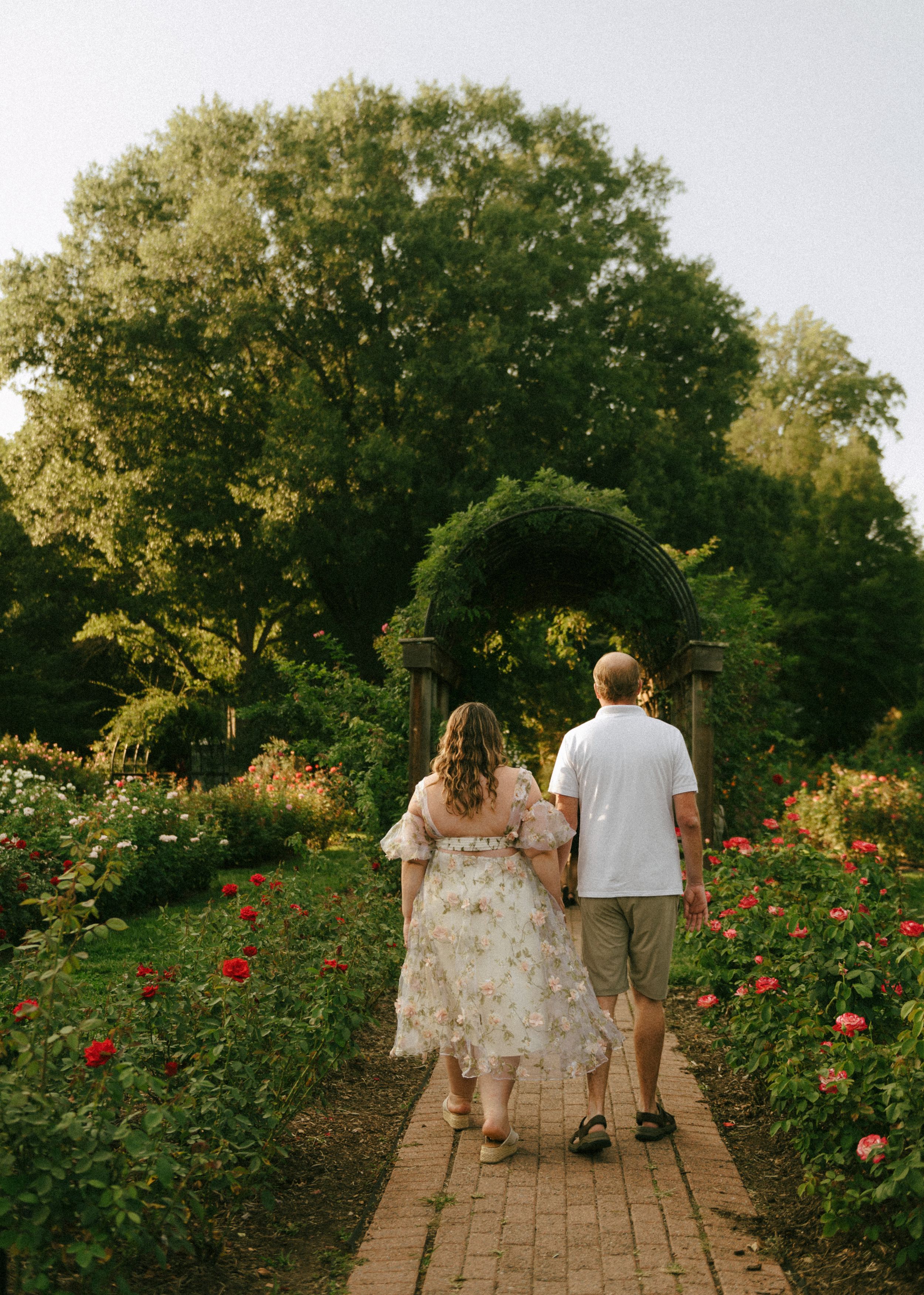 Couple walking hand-in-hand through a lush garden path with red roses and a green archway.