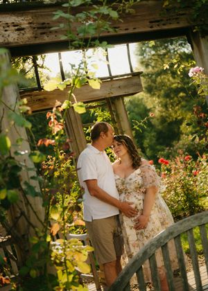 Couple sharing a tender kiss under a rustic garden arbor surrounded by blooming roses, with woman in floral dress cradling baby bump.
