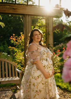 Pregnant woman in floral gown poses in sunlit garden near wooden pergola and roses.