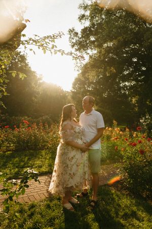 Couple shares a tender moment in a sunlit garden surrounded by colorful roses during a golden hour maternity photoshoot.