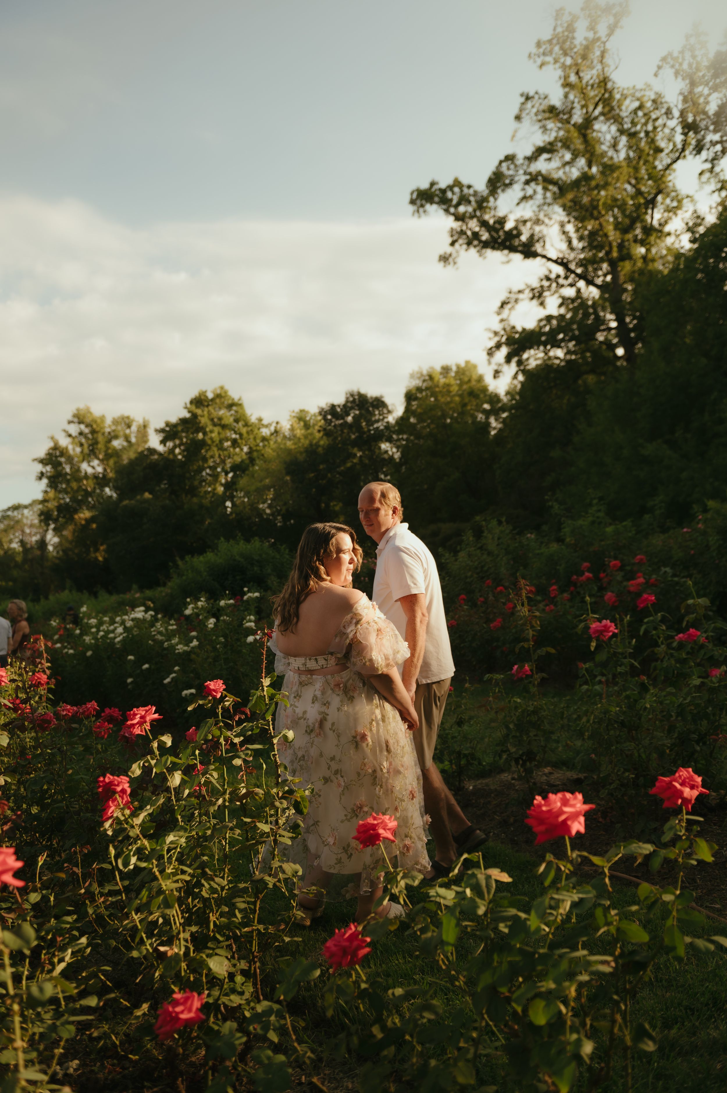 Couple standing among pink roses in a lush garden at golden hour, woman in floral dress, man in white shirt.