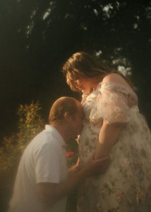 A man kneels before a pregnant woman in a floral dress, tenderly resting his head on her baby bump outdoors.