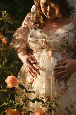 Pregnant woman in floral dress cradling baby bump among blooming peach roses in golden sunlight.