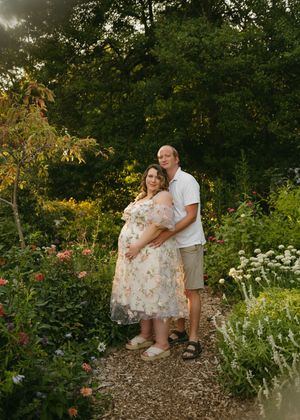 Pregnant couple poses in a lush garden path at golden hour, woman in floral dress cradling baby bump.