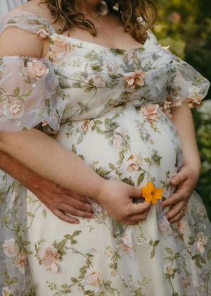 Pregnant woman in floral off-shoulder gown holding a yellow flower, with partner's hands on her baby bump.