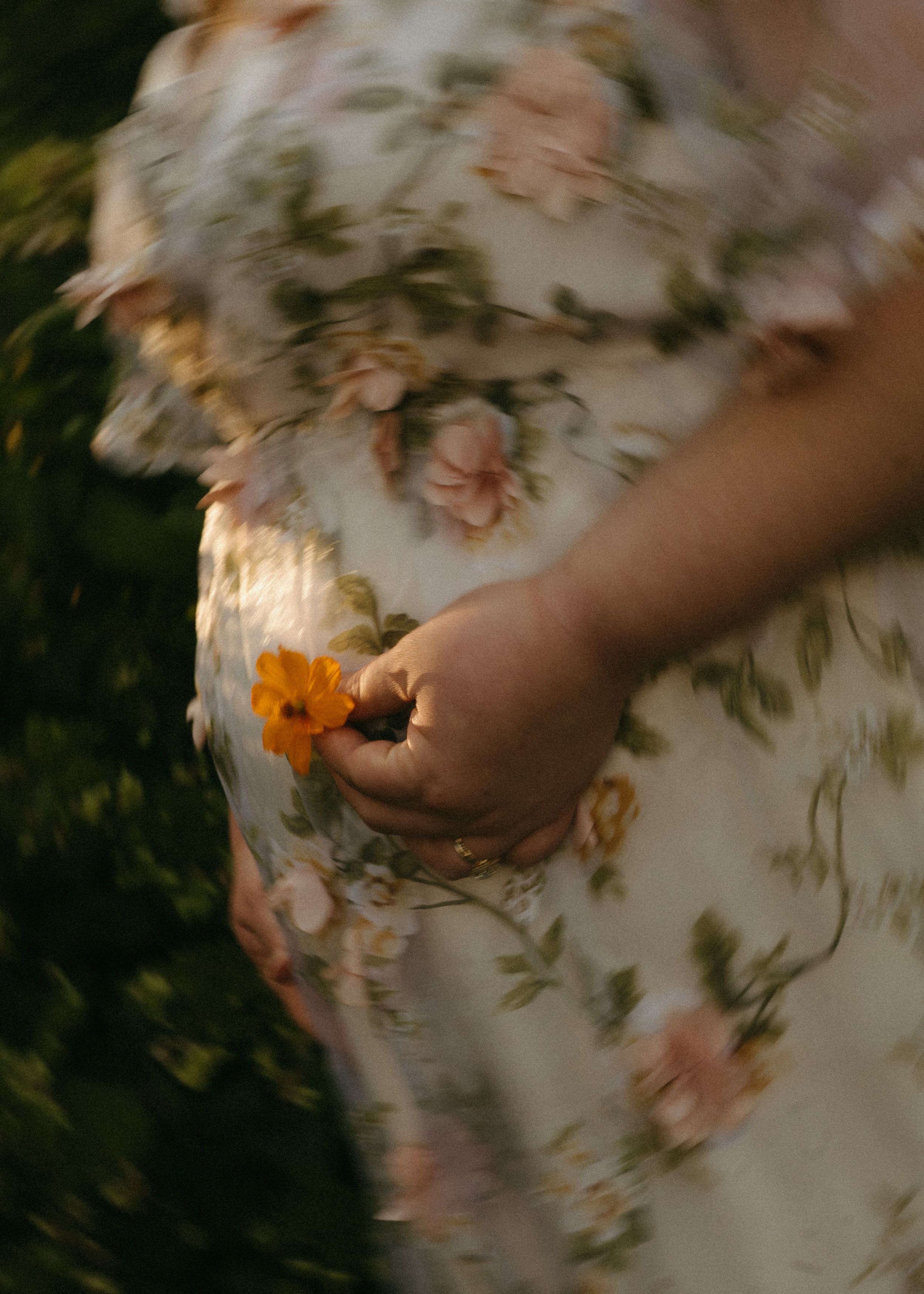 A pregnant woman in a floral dress holds a small orange flower against her baby bump in a garden setting.