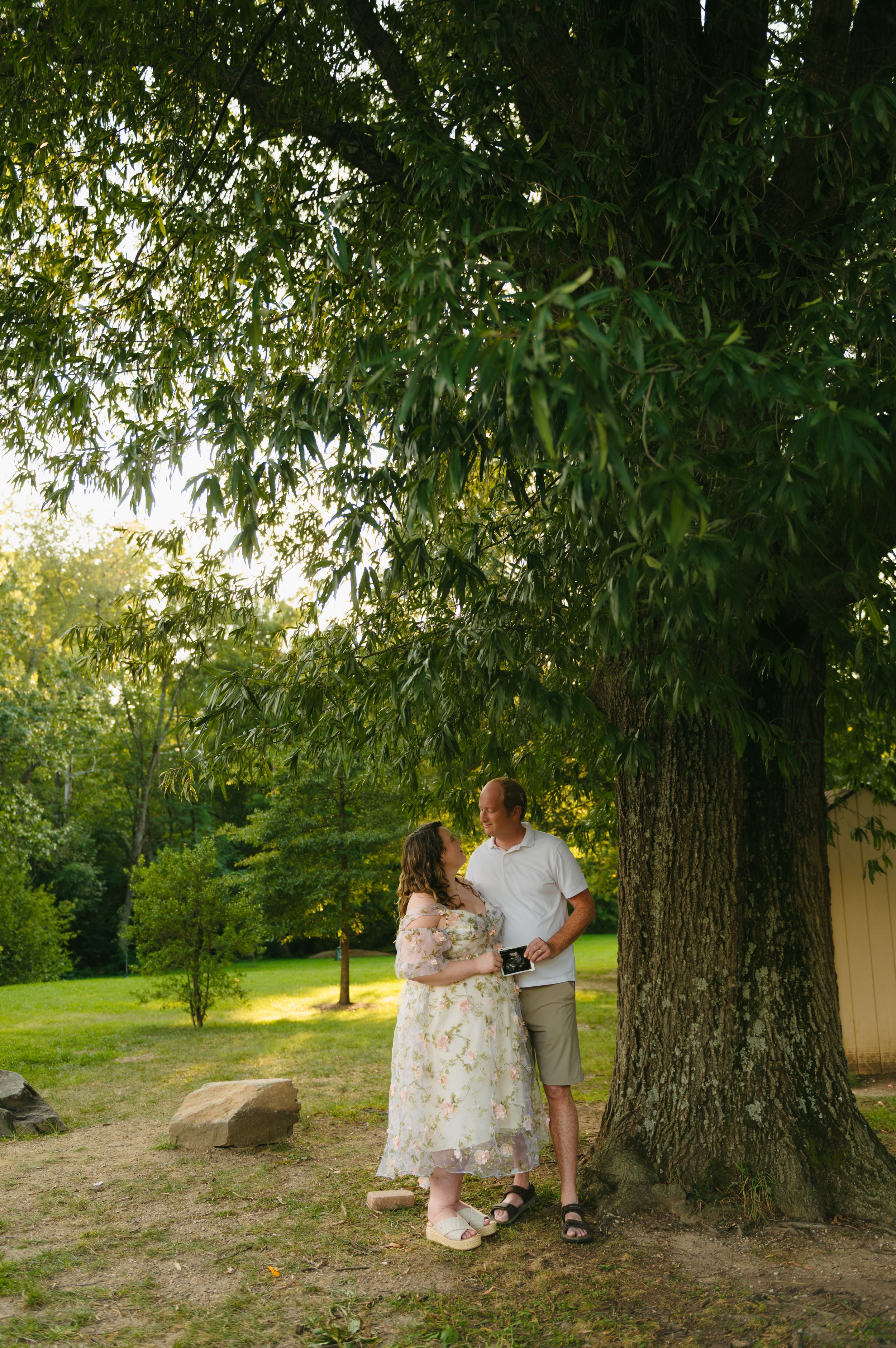 Couple standing under a large tree in a lush green park, woman in floral dress holding ultrasound photo.