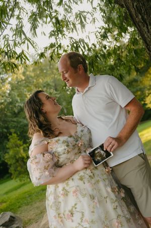 A smiling couple shares a joyful moment outdoors, holding an ultrasound photo, as the woman cradles her baby bump.