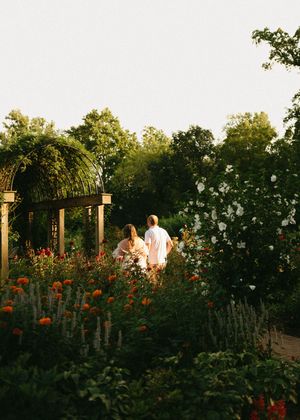 Two people walk through a lush garden filled with orange marigolds and white roses at golden hour.