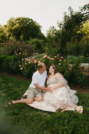 Couple sitting on grass in a rose garden, woman in floral dress and man in white shirt sharing a romantic moment.