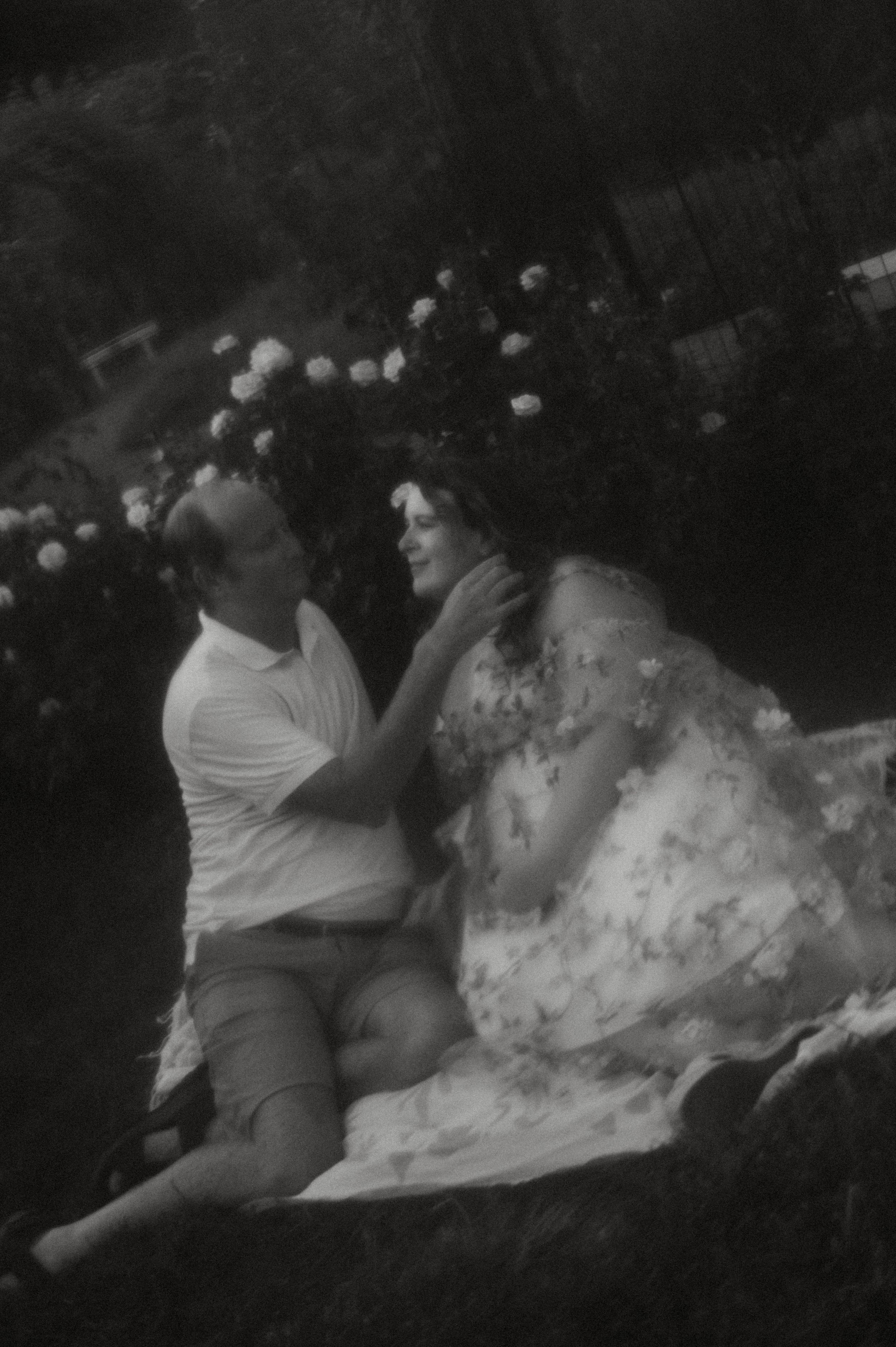 Black and white romantic maternity photo of couple sitting together in a rose garden.