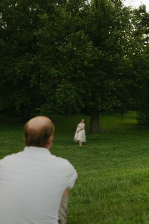 Pregnant woman in floral dress stands among green trees as man watches from foreground.