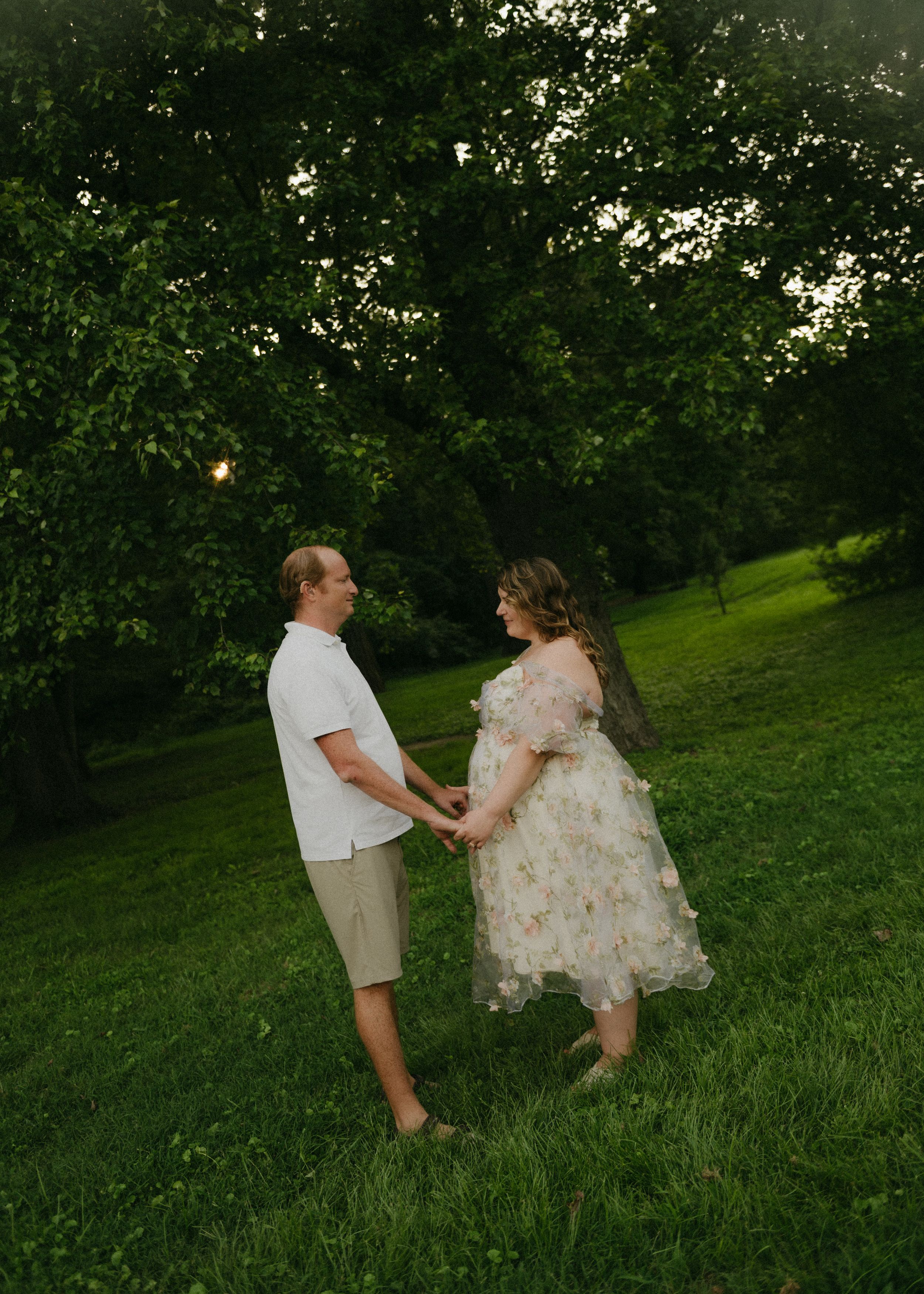 Couple holding hands in a lush green garden at sunset, woman in floral dress showing baby bump.