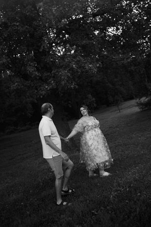 Black and white photo of couple holding hands on grass, woman in floral maternity dress.
