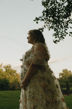 Pregnant woman in floral dress standing peacefully outdoors at dusk under a leafy tree.