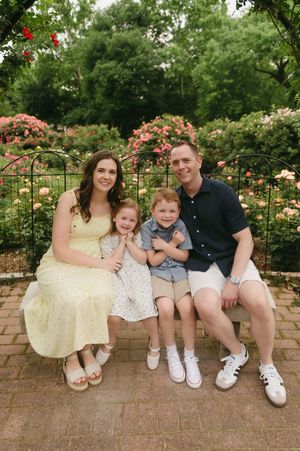 Family of four smiling on a bench surrounded by colorful roses in a lush garden.