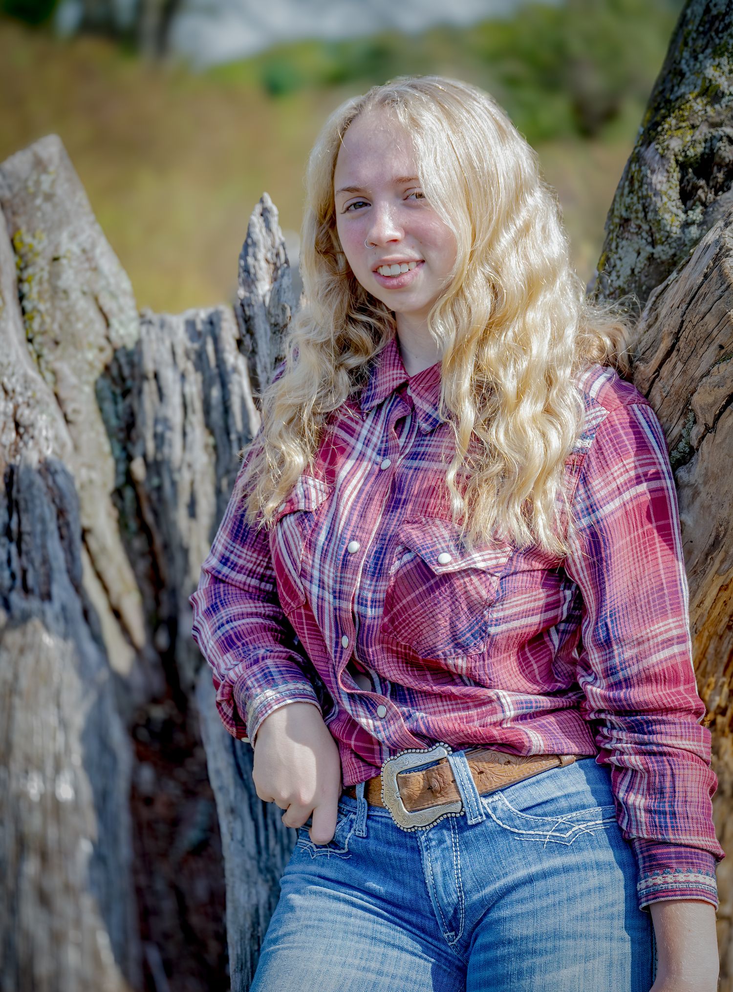 Person in pink plaid shirt and jeans leaning against weathered wood outdoors.